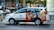 A Vinasun Taxi vehicle is parked on the street, featuring a colorful advertisement on its side promoting instant noodles. The ad displays vibrant images of noodle dishes with text in a foreign language. The vehicle is positioned in front of an urban backdrop with various shops and signs visible, including a restaurant and other businesses.