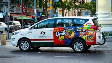 A Vinasun Taxi vehicle is parked on the street, featuring a colorful advertisement on its side promoting instant noodles. The ad displays vibrant images of noodle dishes with text in a foreign language. The vehicle is positioned in front of an urban backdrop with various shops and signs visible, including a restaurant and other businesses.