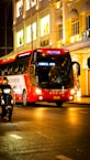 A red and white tour bus travels along a brightly lit city street at night. The buildings in the background feature illuminated windows showcasing mannequins in the display with signs in an Asian script. A person rides a scooter alongside the bus, and the scene is vibrant with an urban nightlife atmosphere.