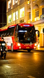 A red and white tour bus travels along a brightly lit city street at night. The buildings in the background feature illuminated windows showcasing mannequins in the display with signs in an Asian script. A person rides a scooter alongside the bus, and the scene is vibrant with an urban nightlife atmosphere.