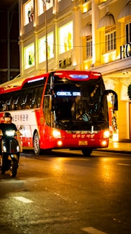 A red and white tour bus travels along a brightly lit city street at night. The buildings in the background feature illuminated windows showcasing mannequins in the display with signs in an Asian script. A person rides a scooter alongside the bus, and the scene is vibrant with an urban nightlife atmosphere.