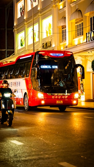 A red and white tour bus travels along a brightly lit city street at night. The buildings in the background feature illuminated windows showcasing mannequins in the display with signs in an Asian script. A person rides a scooter alongside the bus, and the scene is vibrant with an urban nightlife atmosphere.