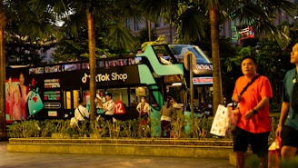 A double-decker bus painted in green and black, featuring advertisements for a TikTok Shop, stands amidst tall palm trees on a bustling city street. People walk by, some carrying shopping bags. The bus is parked, and several individuals are seen near it, possibly waiting or taking photos.