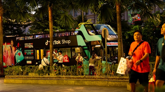 A double-decker bus painted in green and black, featuring advertisements for a TikTok Shop, stands amidst tall palm trees on a bustling city street. People walk by, some carrying shopping bags. The bus is parked, and several individuals are seen near it, possibly waiting or taking photos.