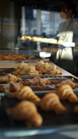 A bakery setting with trays of freshly baked croissants on display, some topped with sliced almonds and cream. A person wearing an apron is visible in the background, managing the baked goods.