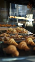 A bakery setting with trays of freshly baked croissants on display, some topped with sliced almonds and cream. A person wearing an apron is visible in the background, managing the baked goods.