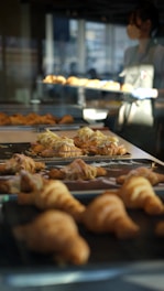 A bakery setting with trays of freshly baked croissants on display, some topped with sliced almonds and cream. A person wearing an apron is visible in the background, managing the baked goods.