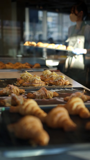 A bakery setting with trays of freshly baked croissants on display, some topped with sliced almonds and cream. A person wearing an apron is visible in the background, managing the baked goods.