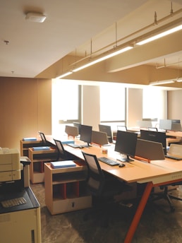 A clean, orderly office space with several computer workstations arranged along the wall. Each desk is equipped with a computer monitor, keyboard, and chair. The room is well-lit with natural light coming in through large windows and overhead fluorescent lighting. There is a printer/copier machine visible in the foreground on the left.