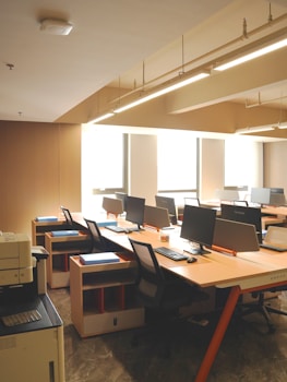 A clean, orderly office space with several computer workstations arranged along the wall. Each desk is equipped with a computer monitor, keyboard, and chair. The room is well-lit with natural light coming in through large windows and overhead fluorescent lighting. There is a printer/copier machine visible in the foreground on the left.