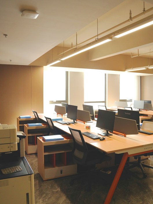 A clean, orderly office space with several computer workstations arranged along the wall. Each desk is equipped with a computer monitor, keyboard, and chair. The room is well-lit with natural light coming in through large windows and overhead fluorescent lighting. There is a printer/copier machine visible in the foreground on the left.