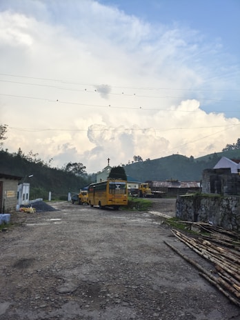 A rural setting featuring yellow school buses parked on a gravel lot surrounded by greenery and hills. There are large, fluffy clouds in the sky, with birds perched on power lines above. A small building and some stacks of bamboo are visible on the right.