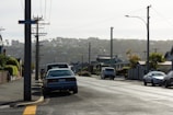 A suburban street scene with parked cars along the side of the road. Power lines and poles line the street, and a street sign labeled 'Dalgety St' is visible on one of the poles. Residential houses with gardens and driveways are on either side, and a hill with more houses is visible in the background under an overcast sky.
