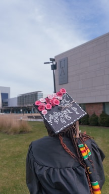 A graduate stands on a grassy campus, wearing a cap decorated with pink and red roses and a personalized message. The gown features colorful African inspired patterns, and the background includes a university building and pathway. The sky is slightly overcast.