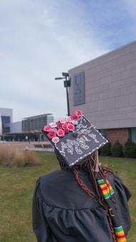 A graduate stands on a grassy campus, wearing a cap decorated with pink and red roses and a personalized message. The gown features colorful African inspired patterns, and the background includes a university building and pathway. The sky is slightly overcast.