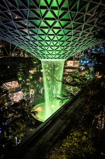 A large indoor waterfall is illuminated with green lighting cascading from an architectural roof structure with a geometric pattern. The surrounding area includes lush greenery and a pathway with small groups of people visible. The ambiance is modern and vibrant, blending nature with architectural elegance.