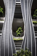 A modern architectural facade with curved, steel-like panels forming wave-like patterns. The design incorporates greenery with several potted trees and shrubs integrated into the structure, creating a harmonious blend of nature and urban design.