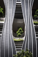 A modern architectural facade with curved, steel-like panels forming wave-like patterns. The design incorporates greenery with several potted trees and shrubs integrated into the structure, creating a harmonious blend of nature and urban design.