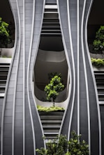 A modern architectural facade with curved, steel-like panels forming wave-like patterns. The design incorporates greenery with several potted trees and shrubs integrated into the structure, creating a harmonious blend of nature and urban design.