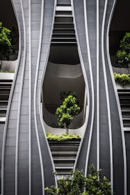 A modern architectural facade with curved, steel-like panels forming wave-like patterns. The design incorporates greenery with several potted trees and shrubs integrated into the structure, creating a harmonious blend of nature and urban design.