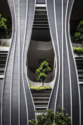 A modern architectural facade with curved, steel-like panels forming wave-like patterns. The design incorporates greenery with several potted trees and shrubs integrated into the structure, creating a harmonious blend of nature and urban design.