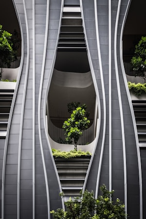 A modern architectural facade with curved, steel-like panels forming wave-like patterns. The design incorporates greenery with several potted trees and shrubs integrated into the structure, creating a harmonious blend of nature and urban design.
