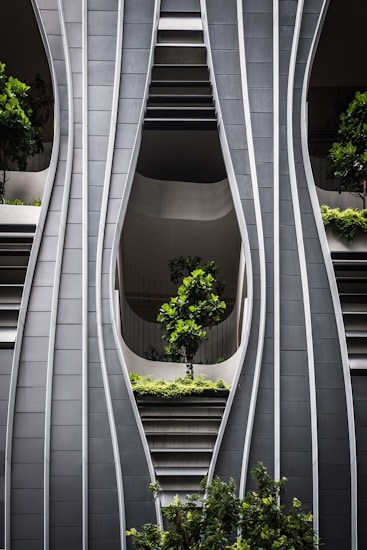 A modern architectural facade with curved, steel-like panels forming wave-like patterns. The design incorporates greenery with several potted trees and shrubs integrated into the structure, creating a harmonious blend of nature and urban design.