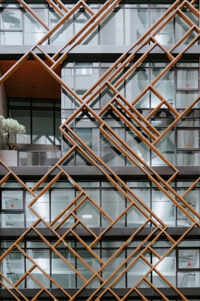 An architectural facade features a geometric pattern created with overlapping diagonal wooden beams framing large glass windows. The design adds depth and texture, with sections of the building's interior visible through the clear glass. A potted plant is placed on a balcony visible in one section of the building.