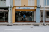 A man is entering a laundry shop with a signboard reading 'Laundry Team' above the entrance. The facade of the building features a mix of yellow and blue tones with adjacent storefronts visible on either side.