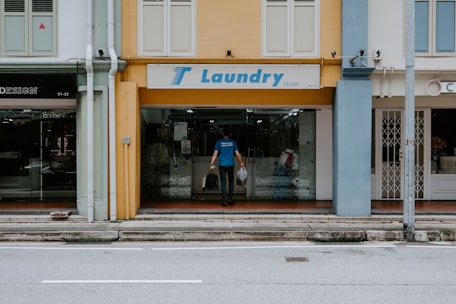 A man is entering a laundry shop with a signboard reading 'Laundry Team' above the entrance. The facade of the building features a mix of yellow and blue tones with adjacent storefronts visible on either side.