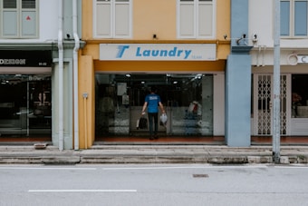A man is entering a laundry shop with a signboard reading 'Laundry Team' above the entrance. The facade of the building features a mix of yellow and blue tones with adjacent storefronts visible on either side.