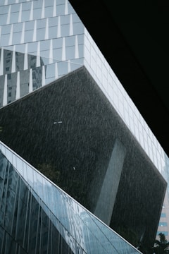 A modern architectural structure with angular shapes and a facade made of glass panels, appearing dark and reflective. Rain is falling heavily, creating streaks across the image and adding a sense of motion. The design is sleek and geometric.