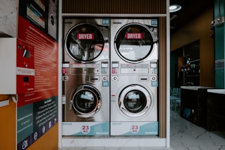 A laundromat interior featuring two stacked commercial washer and dryer machines. The machines have a capacity of 23 kg each. The front view displays control panels with various settings and instructions. The background includes a partially visible reception area with chairs, and there are informational posters mounted on a side wall.