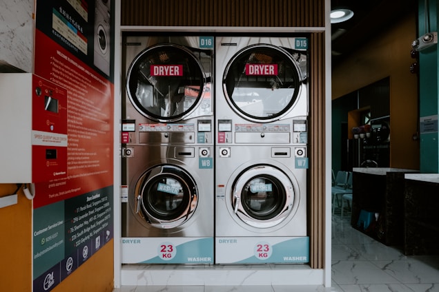 A laundromat interior featuring two stacked commercial washer and dryer machines. The machines have a capacity of 23 kg each. The front view displays control panels with various settings and instructions. The background includes a partially visible reception area with chairs, and there are informational posters mounted on a side wall.