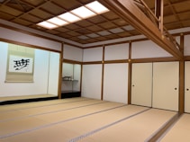 A traditional Japanese room featuring tatami mats covering the floor, sliding doors with a simple circular handle design, and a wooden beamed ceiling. The room has a clean, minimalist aesthetic with a wall scroll displaying calligraphy, adding to the cultural ambiance. A small shelf in the corner holds an ornamental object.