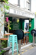 A cozy outdoor cafe with the sign 'Black Sheep & Co' above the entrance. Greenery and pink flowers decorate the entrance area, which features stacked mint green bar stools in front of a dark green wall. Round tabletops are leaning against metal stands while brown wooden bar stools are arranged next to the tables inside. A chalkboard with inspirational text adds character to the scene.
