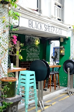 A cozy outdoor cafe with the sign 'Black Sheep & Co' above the entrance. Greenery and pink flowers decorate the entrance area, which features stacked mint green bar stools in front of a dark green wall. Round tabletops are leaning against metal stands while brown wooden bar stools are arranged next to the tables inside. A chalkboard with inspirational text adds character to the scene.