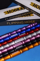The image features several metallic hookah hoses in various colors, including purple, blue, red, silver, pink, black, and gold. These hoses are arranged diagonally across a blue surface. In the background, there are boxed packaging with the words 'Flipping Hose' and 'Logic Hookah' partially visible.