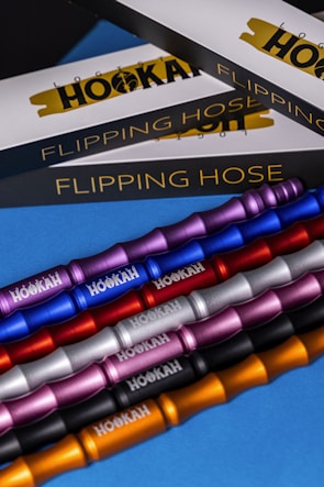 The image features several metallic hookah hoses in various colors, including purple, blue, red, silver, pink, black, and gold. These hoses are arranged diagonally across a blue surface. In the background, there are boxed packaging with the words 'Flipping Hose' and 'Logic Hookah' partially visible.