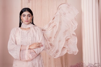 A woman stands confidently with her arms crossed, wearing an elegant, pastel pink traditional outfit with intricate embroidery. Her long, dark hair is straightened, and she accessorizes with delicate earrings. A sheer scarf flows beside her, adding to the poised and serene atmosphere against a soft beige backdrop with draped fabric and a column.