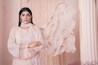 A woman stands confidently with her arms crossed, wearing an elegant, pastel pink traditional outfit with intricate embroidery. Her long, dark hair is straightened, and she accessorizes with delicate earrings. A sheer scarf flows beside her, adding to the poised and serene atmosphere against a soft beige backdrop with draped fabric and a column.