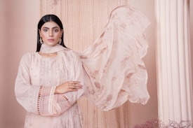 A woman stands confidently with her arms crossed, wearing an elegant, pastel pink traditional outfit with intricate embroidery. Her long, dark hair is straightened, and she accessorizes with delicate earrings. A sheer scarf flows beside her, adding to the poised and serene atmosphere against a soft beige backdrop with draped fabric and a column.