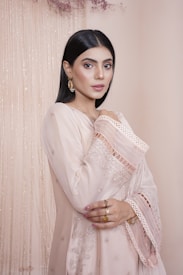 A woman with sleek, dark hair stands against a soft, beige backdrop. She is wearing an elegant, traditional dress with intricate embroidery and lace details. Her accessories include ornate earrings and rings. The setting is enhanced by delicate hanging decor in subdued tones.