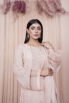 A woman stands elegantly, wearing a detailed beige dress with intricate embroidery and lace patterns. Her hair is straight and she is adorned with dangling gold earrings. The background is decorated with vertical shimmering strings and dried flowers at the top.