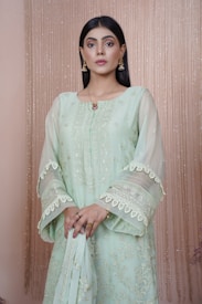 A woman stands elegantly against a neutral backdrop with vertical shimmering lines. She wears a pale green embroidered outfit adorned with intricate patterns and lace detailing, complemented by golden earrings. Her hands hold onto a matching fabric, and her hair is sleek and straight.