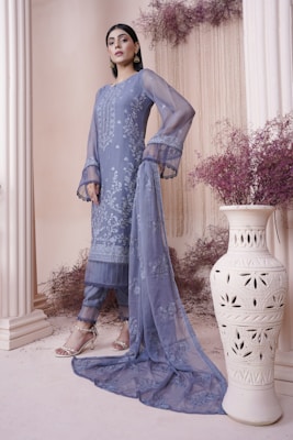A woman stands gracefully in an elegant outfit composed of a long, embroidered sheer fabric with a matching scarf. The ensemble features intricate floral patterns in shades of blue. She is wearing strappy sandals, and the setting is enhanced by dried purple flowers and a carved white vase, giving a sophisticated and artistic ambiance.
