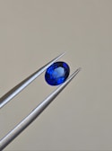 A pair of metallic tweezers delicately holds a small, oval-cut blue gemstone against a neutral background. The gemstone displays a vibrant blue color with facets that catch the light.
