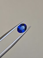 A pair of metallic tweezers delicately holds a small, oval-cut blue gemstone against a neutral background. The gemstone displays a vibrant blue color with facets that catch the light.