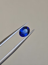 A pair of metallic tweezers delicately holds a small, oval-cut blue gemstone against a neutral background. The gemstone displays a vibrant blue color with facets that catch the light.
