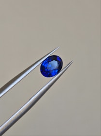A pair of metallic tweezers delicately holds a small, oval-cut blue gemstone against a neutral background. The gemstone displays a vibrant blue color with facets that catch the light.